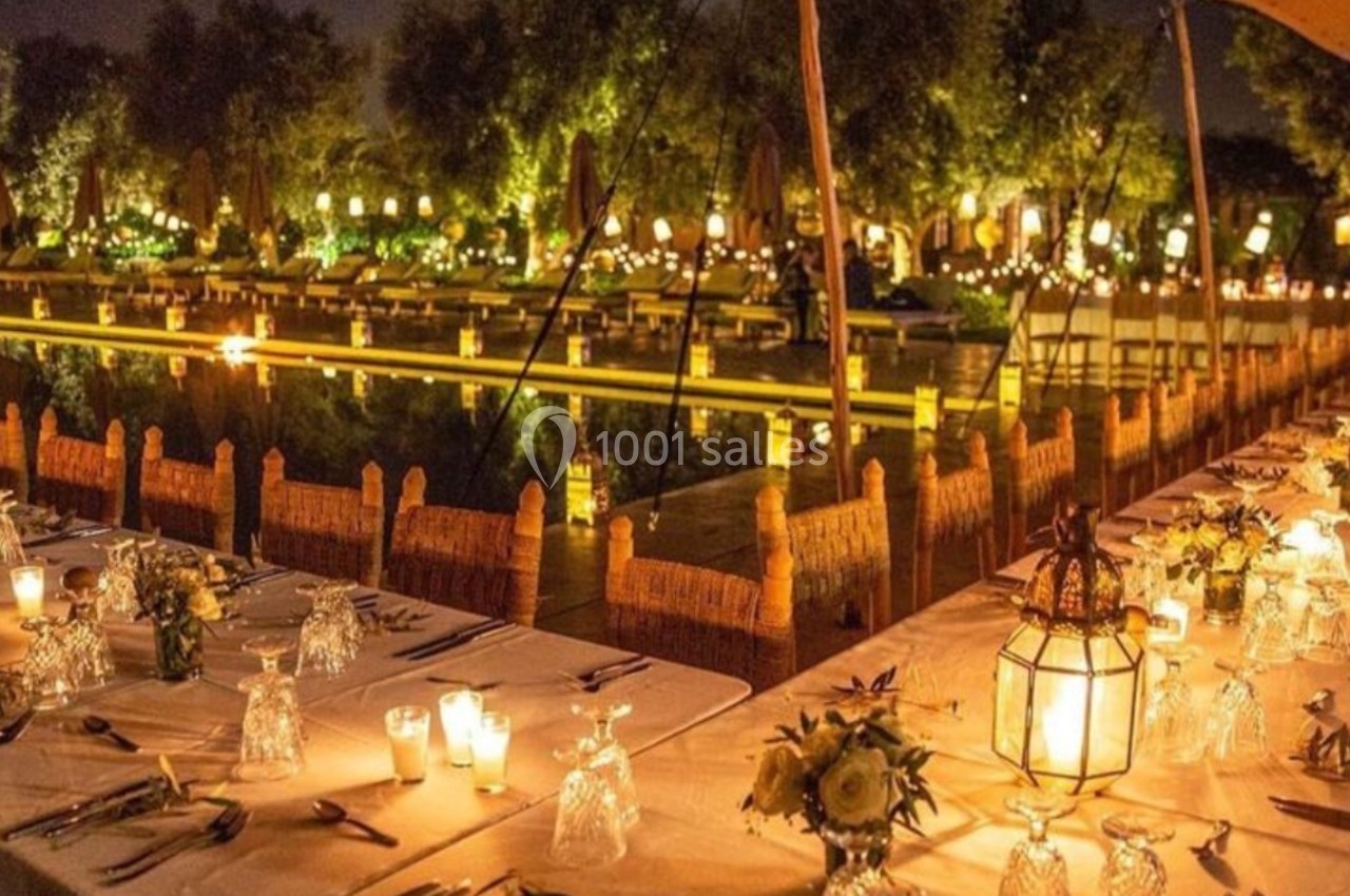 Tables élégamment dressées avec bougies et lanternes, face à un jardin illuminé et un plan d'eau, de nuit.