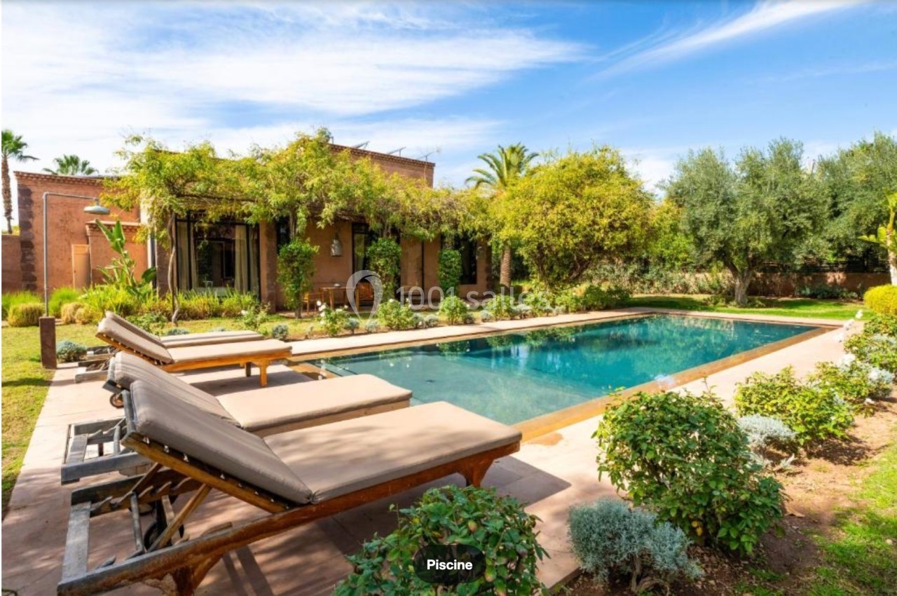 Piscine extérieure entourée de chaises longues, végétation luxuriante et maison en arrière-plan.