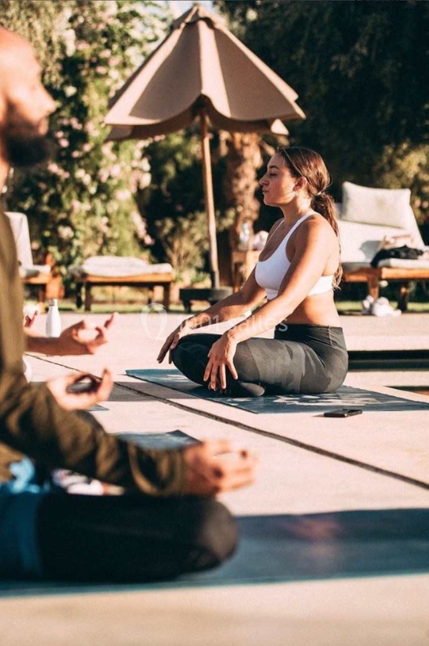 Deux personnes méditent en plein air sur des tapis, près d'une piscine et entourées de végétation.