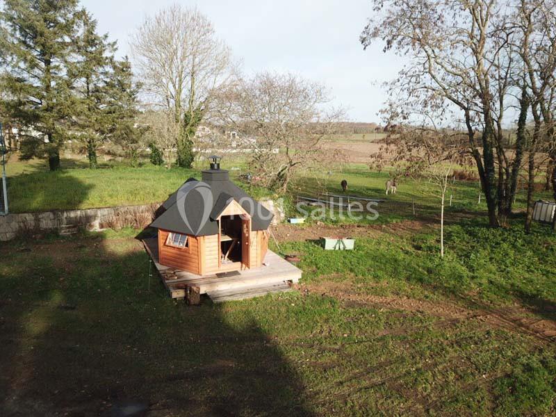Cabane en bois avec toit sombre, située dans un jardin verdoyant entouré d'arbres et de champs.