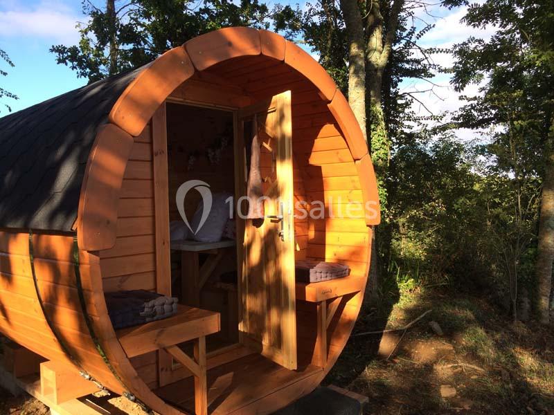 Cabane en bois en forme de tonneau avec porte ouverte, située en pleine nature sous des arbres.