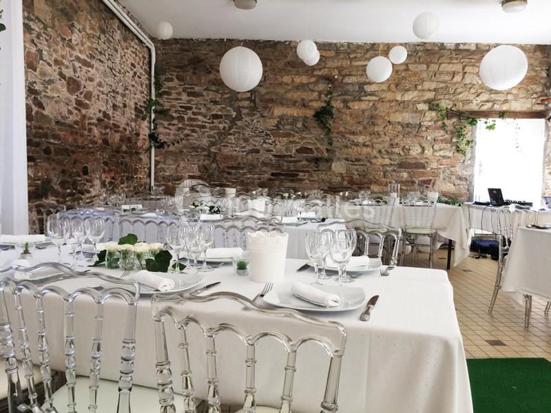 Salle de réception décorée avec des tables dressées, nappes blanches, chaises transparentes et lanternes suspendues.