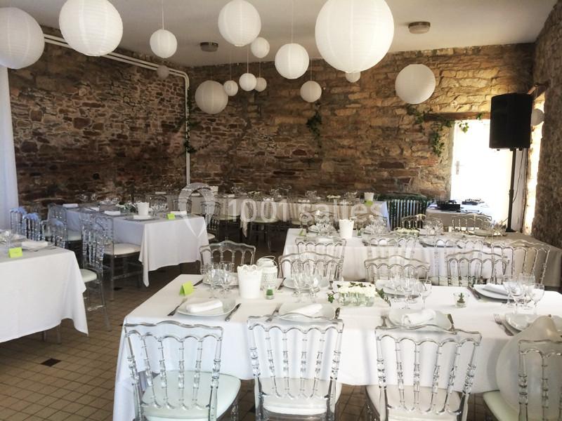 Salle de réception en pierre avec tables dressées, chaises transparentes et lanternes blanches suspendues.