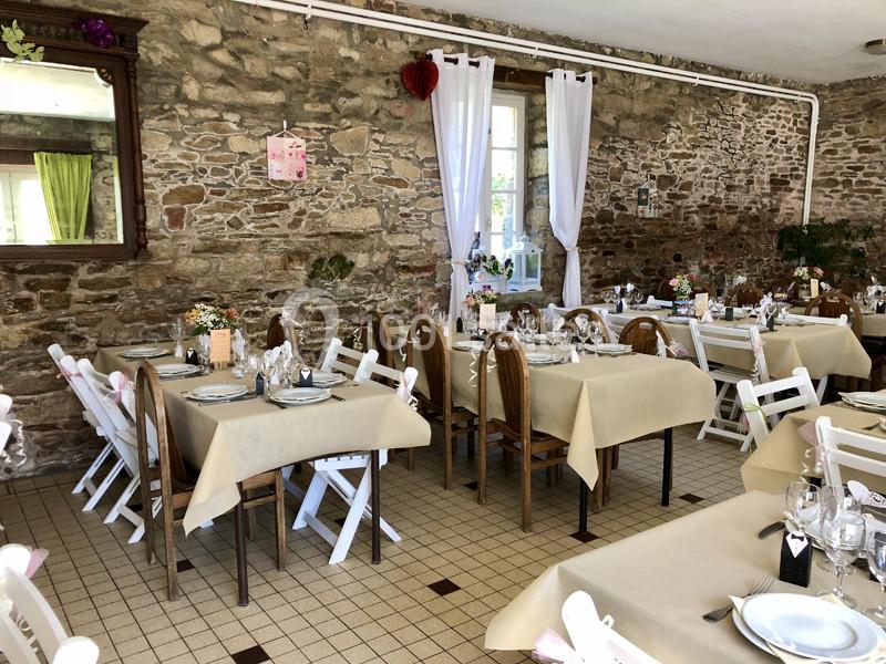 Salle de restaurant avec murs en pierre, tables dressées avec nappes beige et chaises en bois ou blanches pliantes.