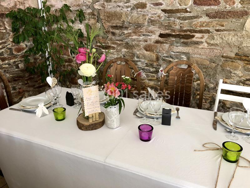 Table décorée pour un repas, avec nappes blanches, fleurs colorées, bougies et vaisselle élégante devant un mur en pierre.