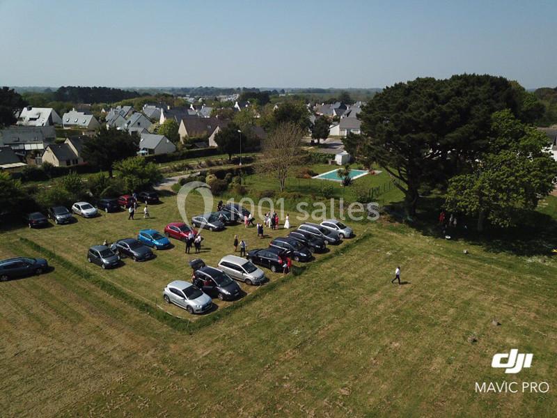 Vue aérienne d'un parking improvisé sur une pelouse, entouré de maisons et d'arbres, avec des personnes rassemblées.