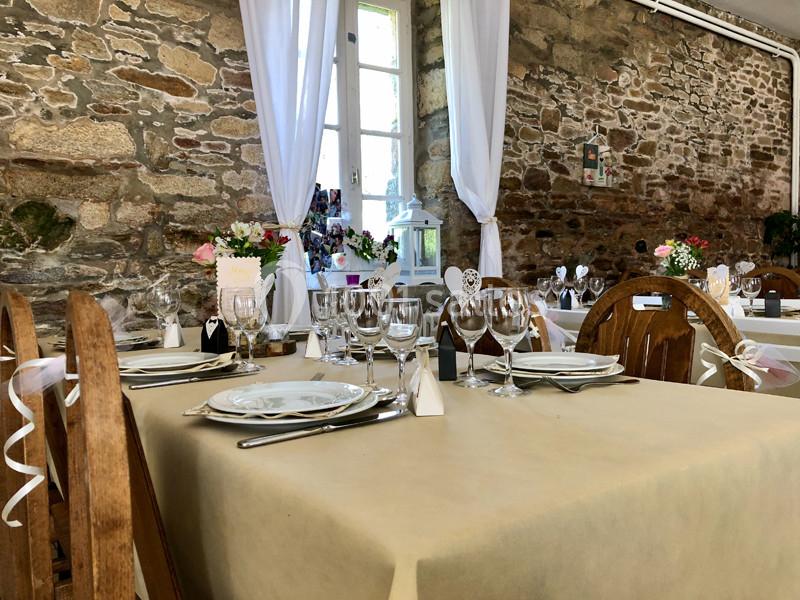 Salle de réception avec tables dressées, nappes beige, vaisselle élégante et murs en pierre apparente.