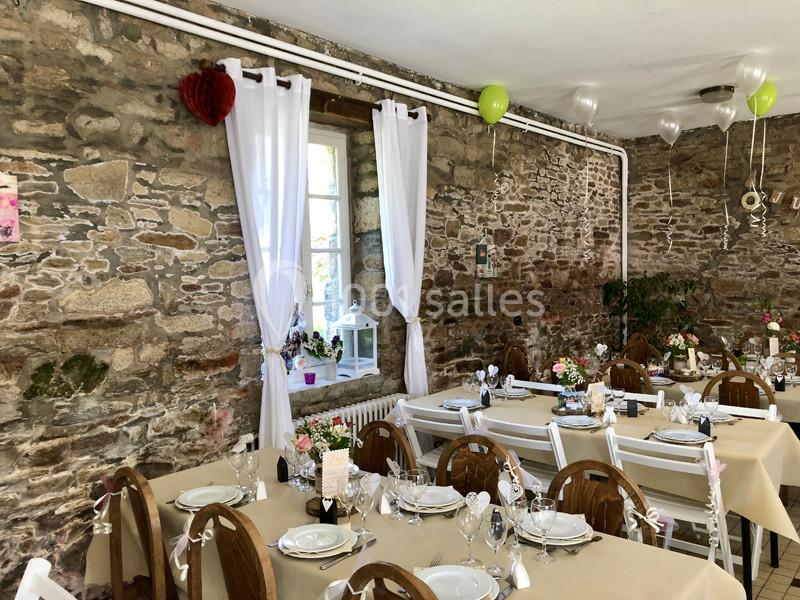 Salle décorée pour un événement, avec des tables dressées, des chaises en bois et des murs en pierre apparente.