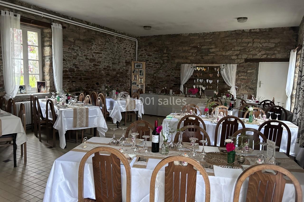Salle de réception décorée avec des tables dressées, nappes blanches, chaises en bois et murs en pierre apparente.