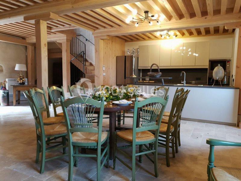 Salle à manger lumineuse avec table ronde entourée de chaises vertes, cuisine ouverte et poutres apparentes en bois.