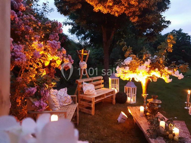 Espace extérieur aménagé avec des bancs en bois, lanternes et bougies, entouré de fleurs et éclairé à la tombée de la nuit.