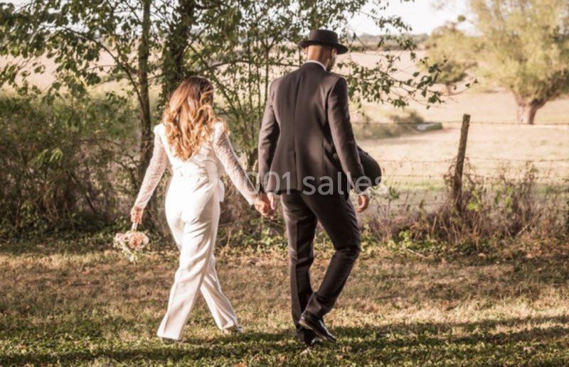 Un couple élégant marche main dans la main dans un champ ensoleillé, entouré d'arbres et de verdure.