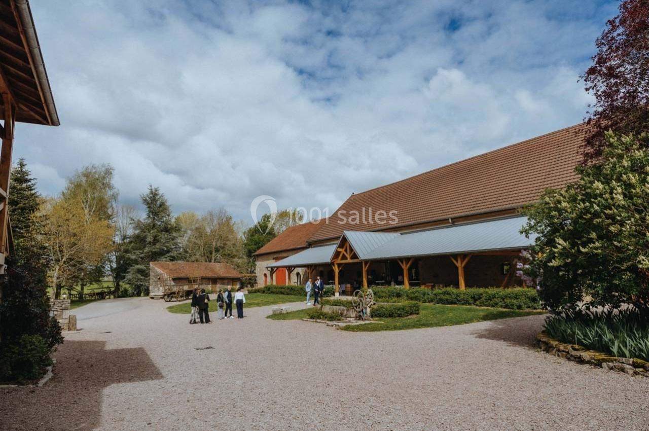 Cour d'une ferme avec bâtiments en pierre et en bois, quelques arbres, et un petit groupe de personnes discutant.