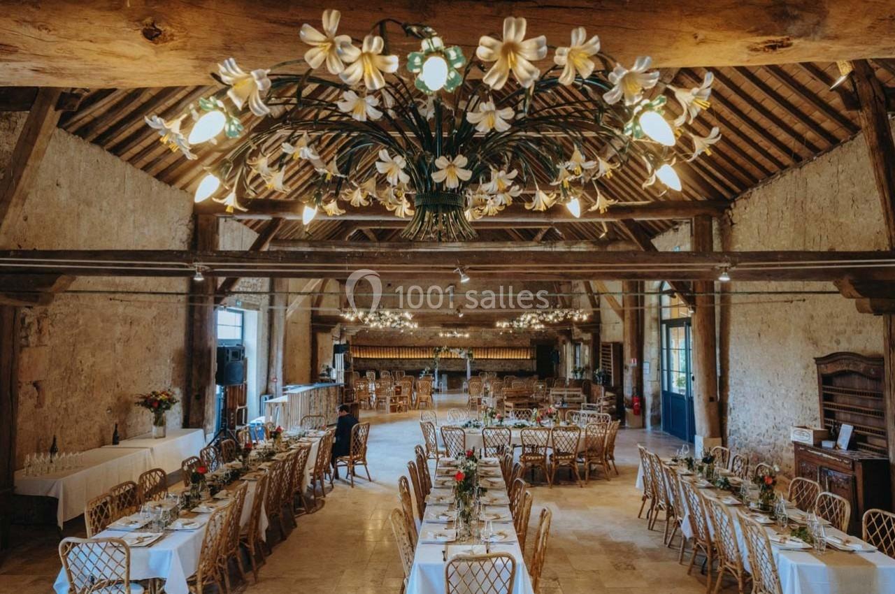 Salle de réception rustique avec tables dressées, chaises en bois et lustres ornés de fleurs blanches.