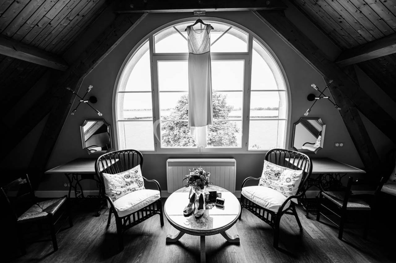 Robe de mariée suspendue devant une grande fenêtre cintrée dans une pièce aménagée avec des fauteuils et une table basse.