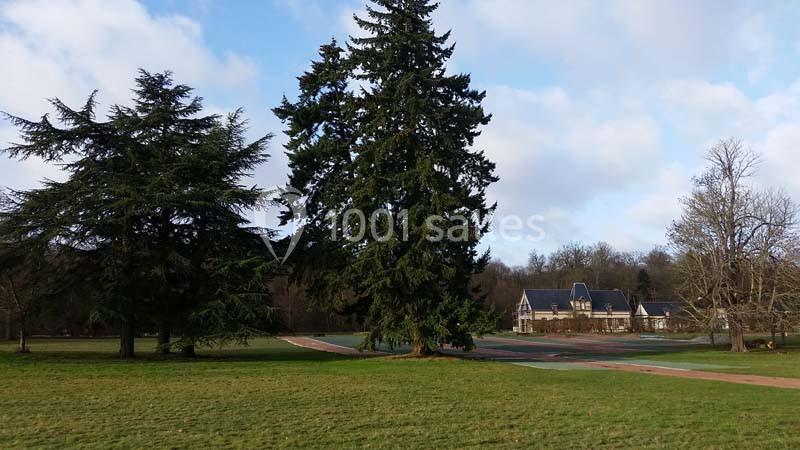 Grand parc avec pelouse, allées pavées, arbres imposants et une maison en arrière-plan sous un ciel partiellement nuageux.