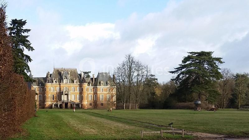 Château en briques rouges entouré de pelouses, d'arbres et d'un ciel partiellement nuageux.