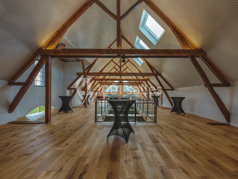 Grande pièce sous combles avec poutres apparentes, parquet en bois clair, tables hautes et puits de lumière.