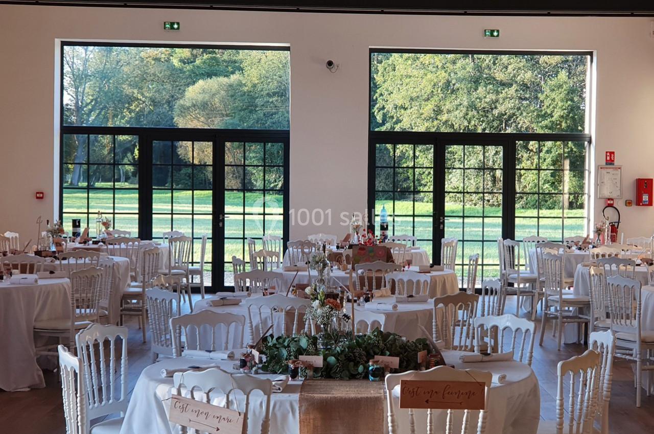 Salle de réception lumineuse avec tables décorées, grandes baies vitrées donnant sur un parc verdoyant.