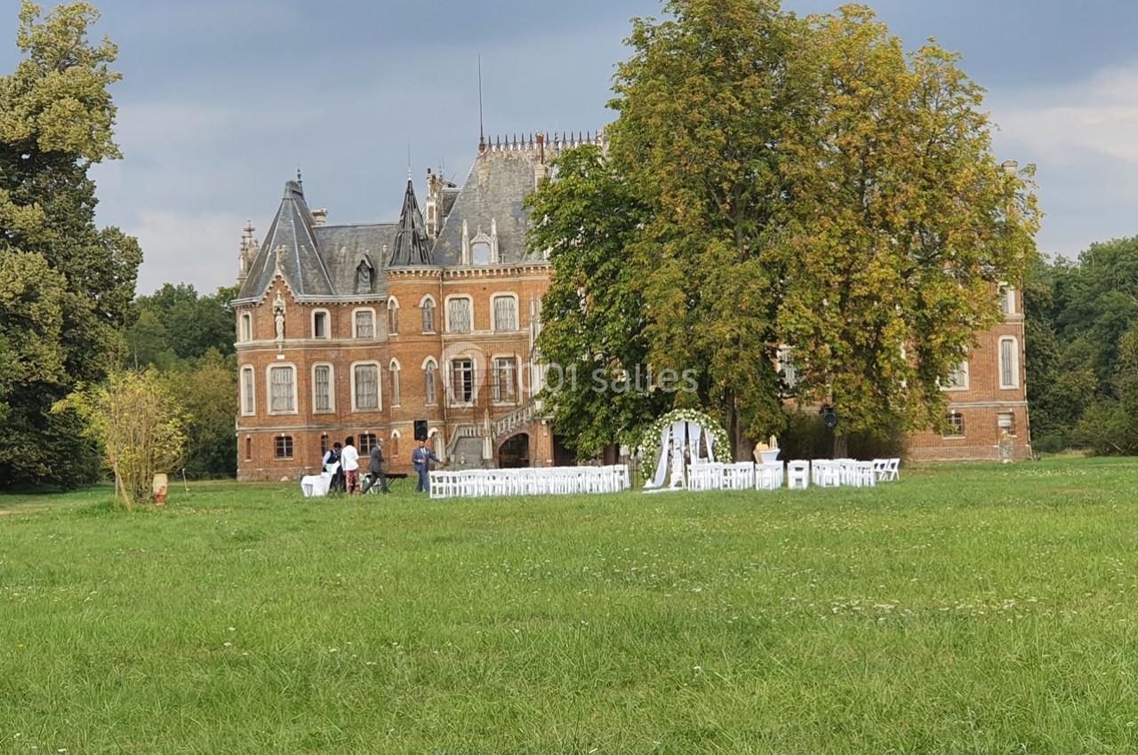 Château en briques entouré de verdure avec des chaises blanches disposées pour une cérémonie en plein air.