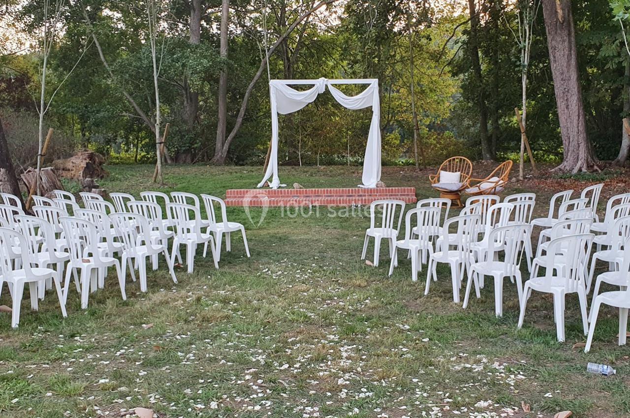 Chaises blanches disposées en rangées devant une arche décorée de tissu blanc dans un cadre extérieur boisé.