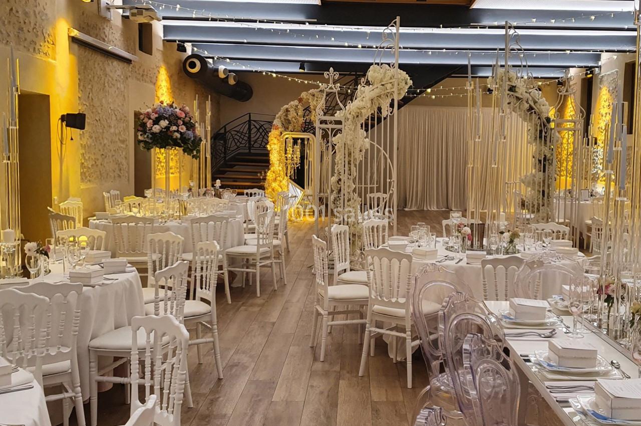 Salle de réception décorée pour un mariage, avec tables dressées, chaises blanches et arches ornées de fleurs.