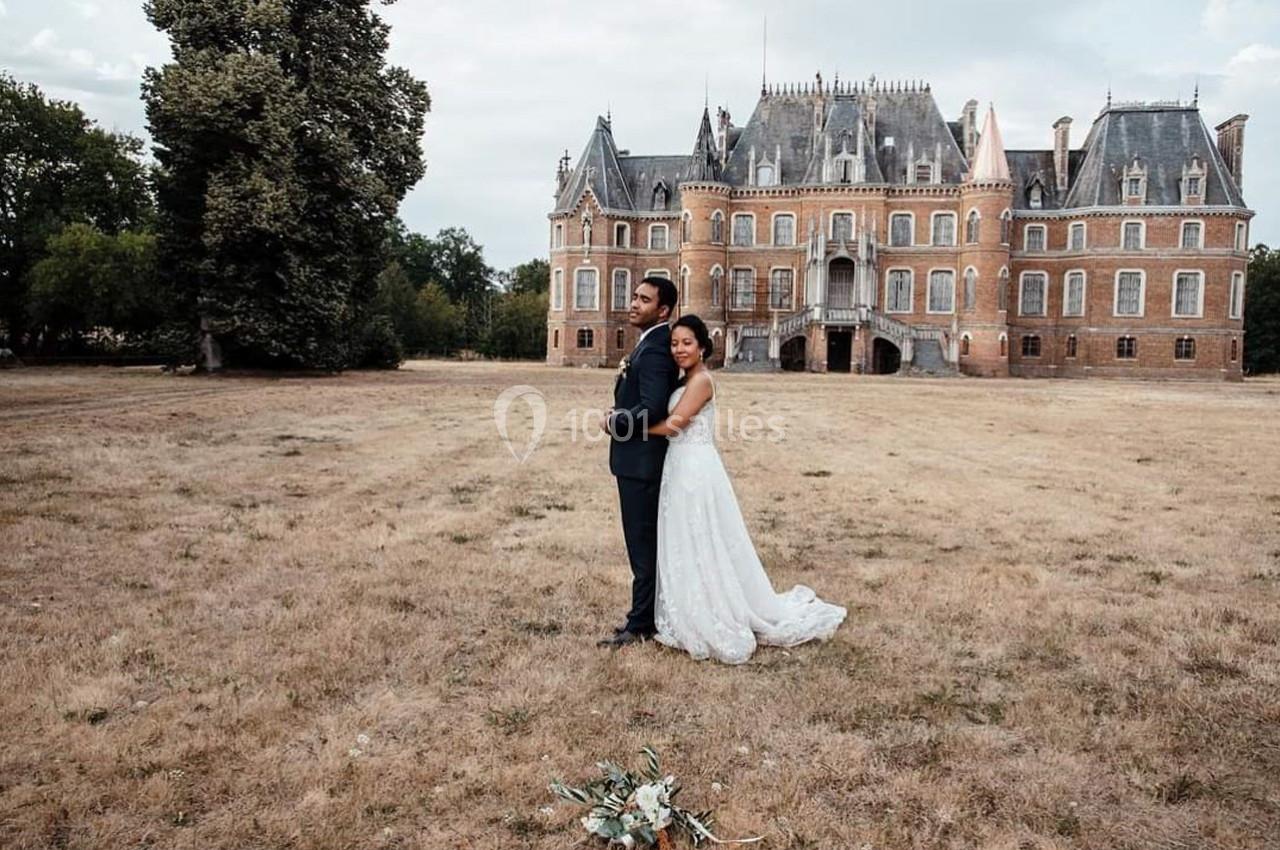 Un couple en tenue de mariage pose devant un château entouré d'une pelouse et d'arbres.