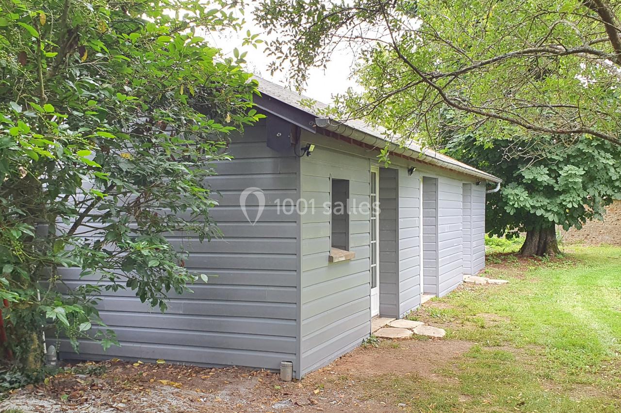 Abri de jardin en bois gris clair avec fenêtres, entouré d'arbres et d'une pelouse verdoyante.