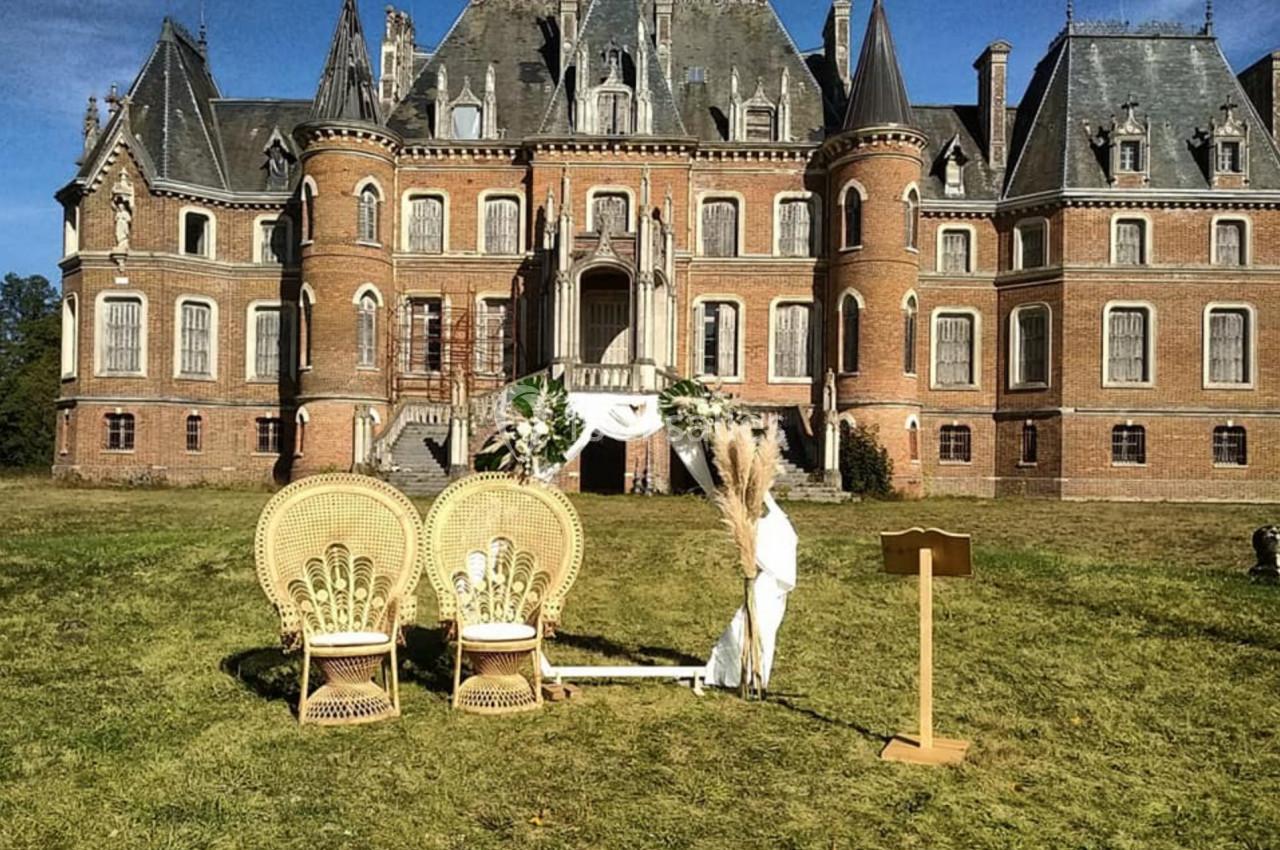 Deux fauteuils en osier et une arche décorée devant un château en briques rouges sous un ciel bleu.