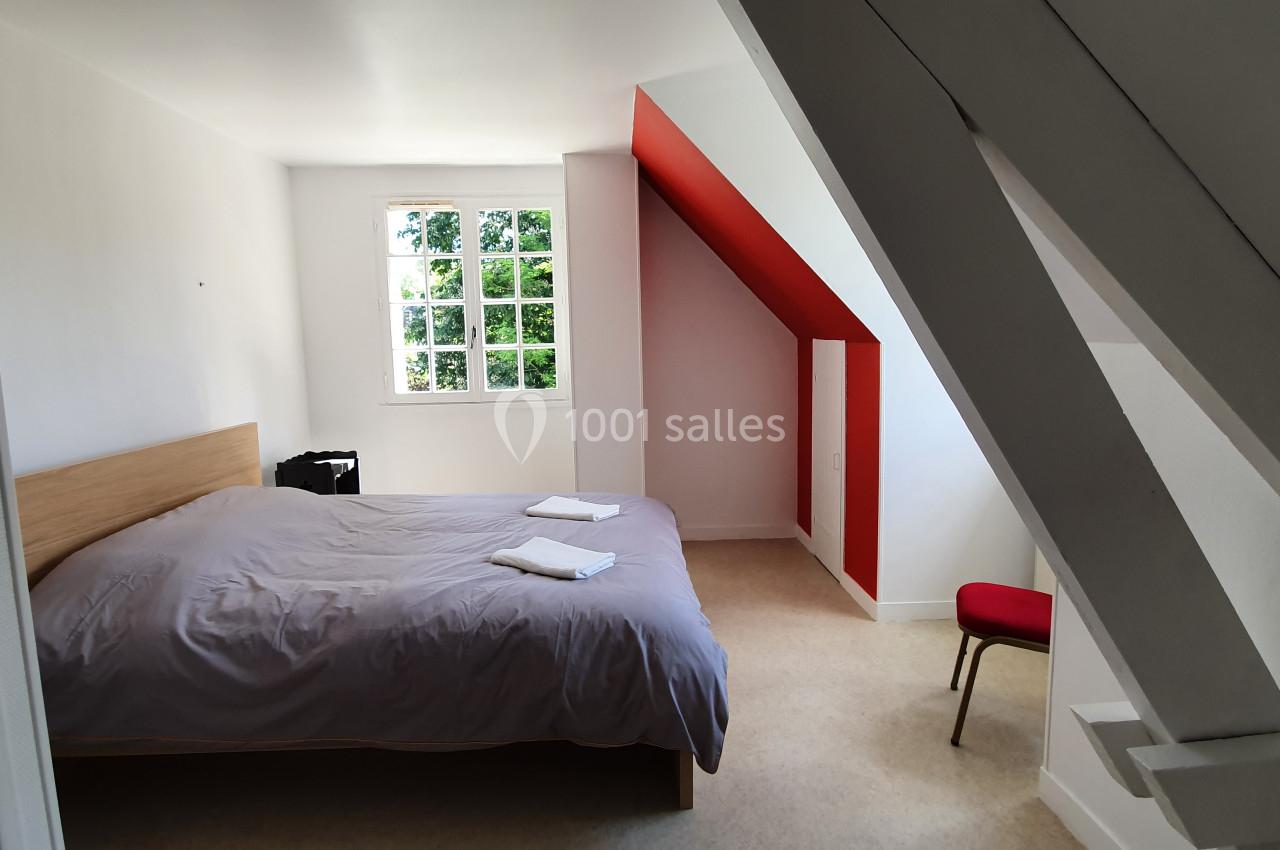 Chambre mansardée lumineuse avec un lit double, draps gris, chaise rouge et fenêtre donnant sur un espace verdoyant.