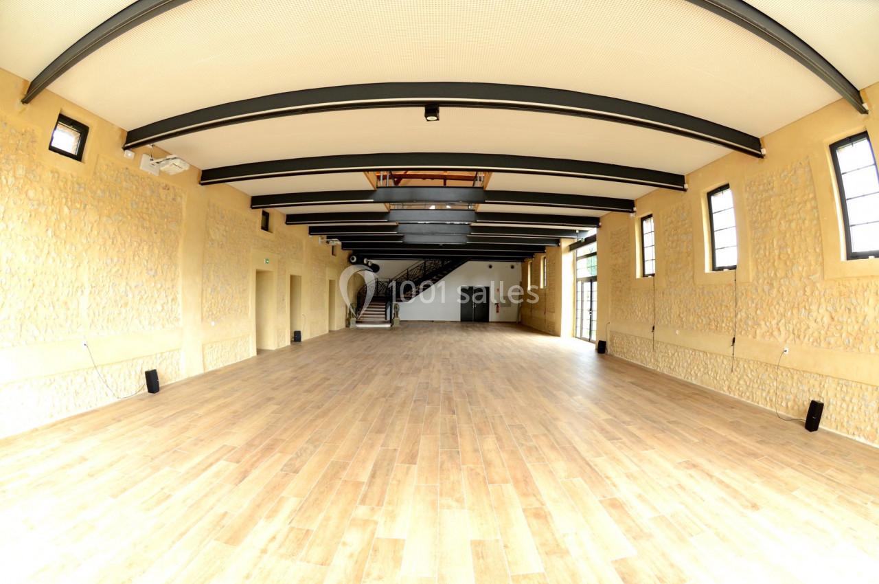 Salle spacieuse avec parquet en bois, murs en pierre et poutres apparentes, éclairée par des fenêtres latérales.