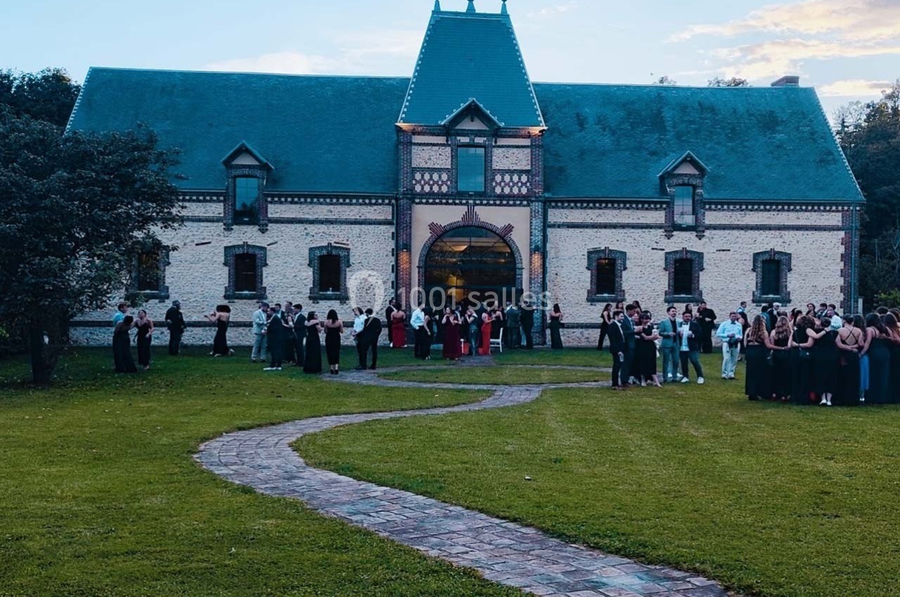 Un groupe de personnes rassemblées devant un grand bâtiment en briques avec un toit en ardoise, entouré de pelouse.