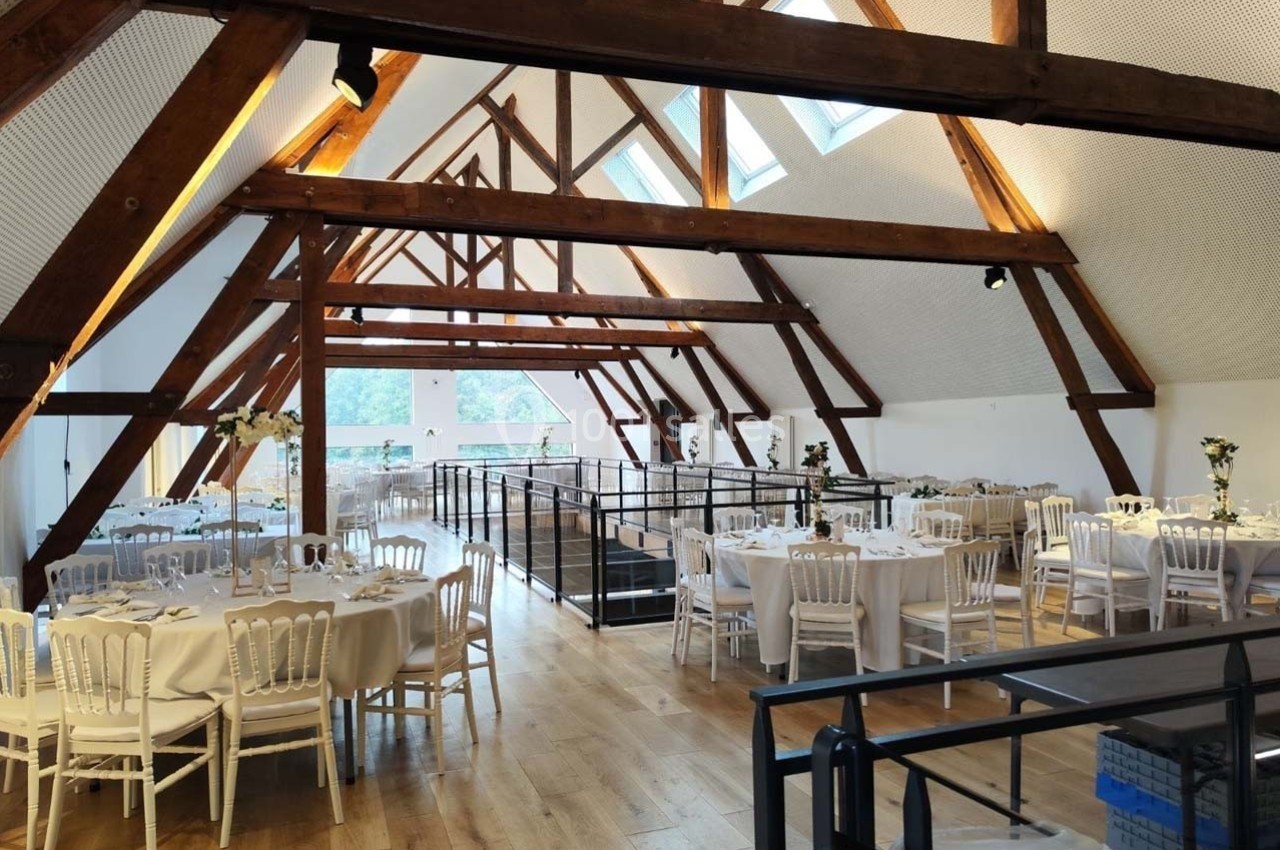 Salle lumineuse avec poutres apparentes, tables rondes dressées pour un événement, parquet en bois clair et baies vitrées.