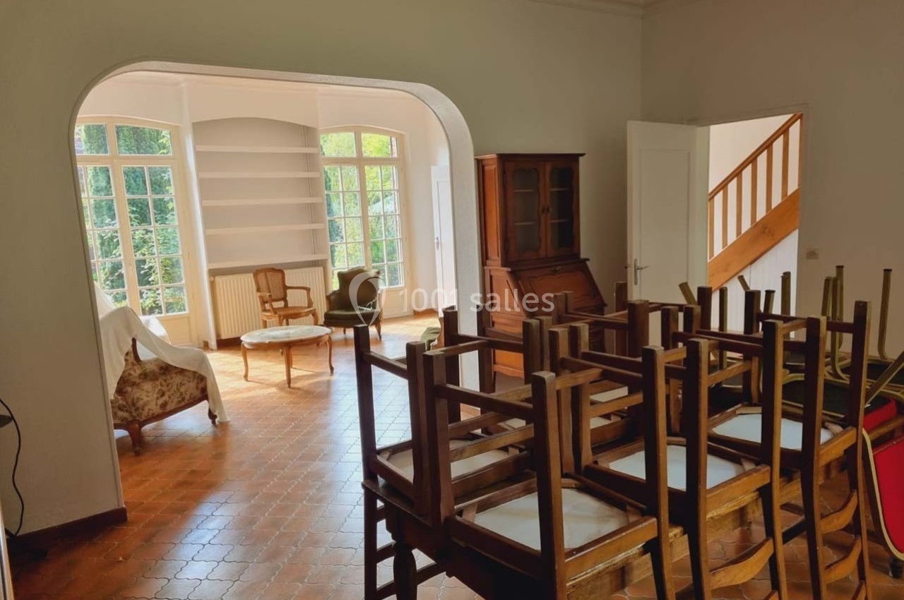 Salle lumineuse avec chaises empilées, sol en terre cuite, meubles en bois et grandes fenêtres donnant sur un jardin.