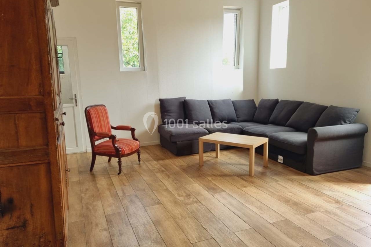 Salon lumineux avec parquet, canapé d'angle gris, fauteuil rouge et table basse en bois clair.