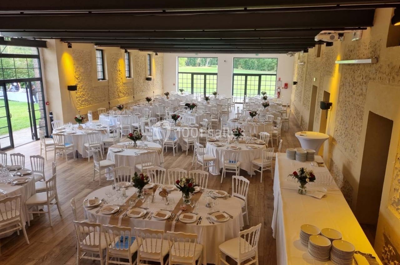 Salle de réception lumineuse avec tables rondes dressées, chaises blanches et décorations florales, vue sur un jardin.