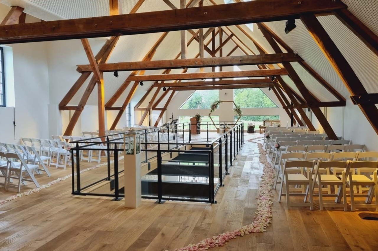 Salle lumineuse avec poutres apparentes, chaises blanches alignées et allée décorée de pétales de fleurs.