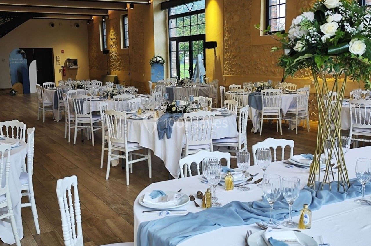 Salle de réception élégamment décorée avec des tables rondes, nappes blanches, chemins de table bleus et compositions…