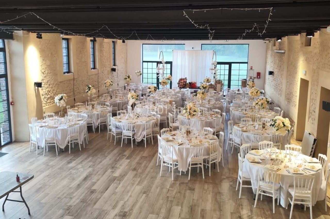 Salle de réception décorée avec des tables rondes dressées, chaises blanches et arrangements floraux élégants.