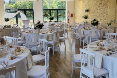 Salle de réception décorée avec des tables rondes, nappes blanches, chaises blanches et centres de table fleuris.