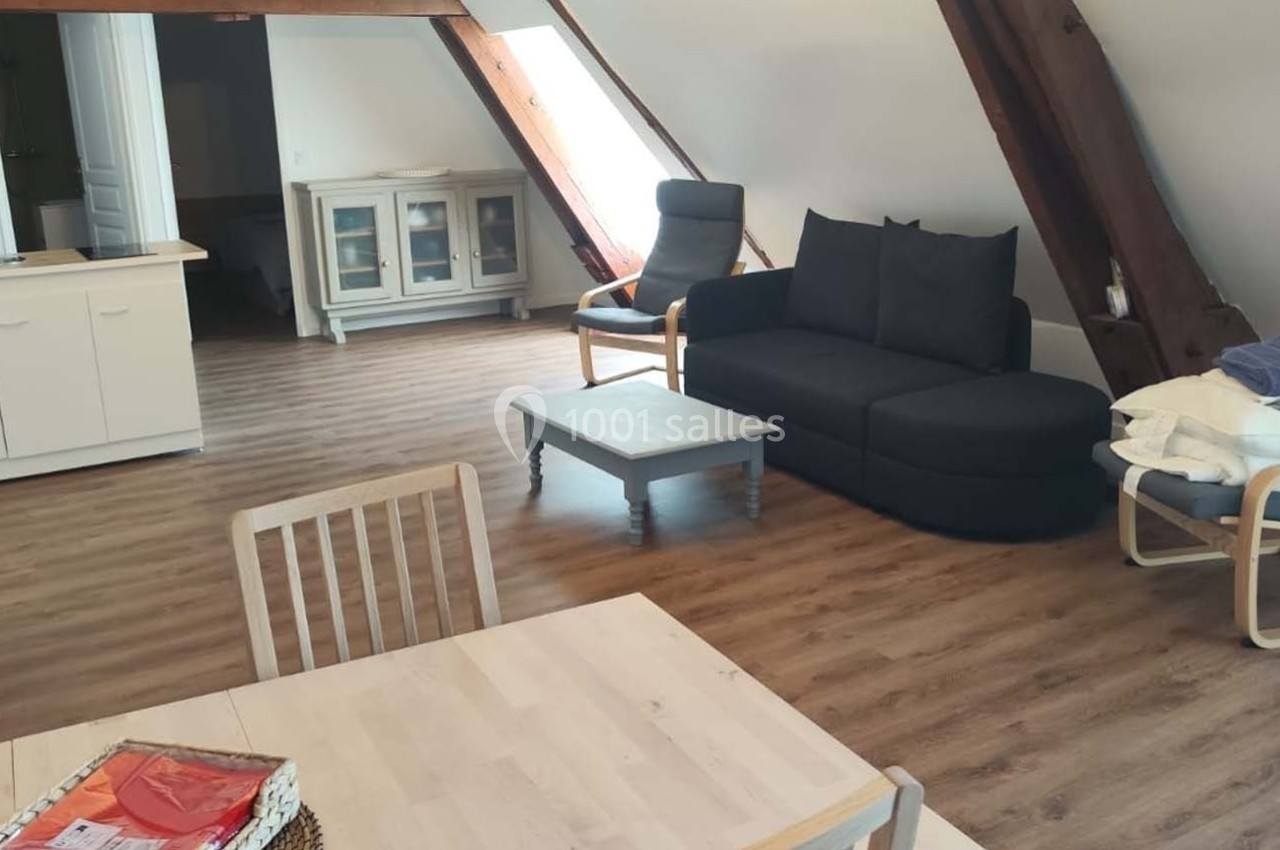 Salon mansardé avec parquet, canapé noir, fauteuil, table basse, table à manger et cuisine ouverte en arrière-plan.