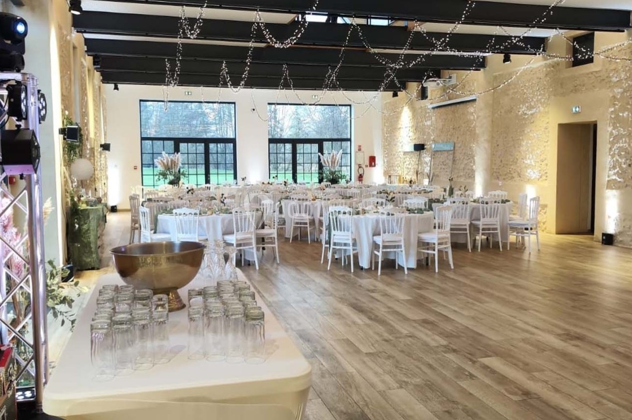 Salle de réception lumineuse avec tables dressées, chaises blanches et décorations suspendues, vue sur un extérieur…