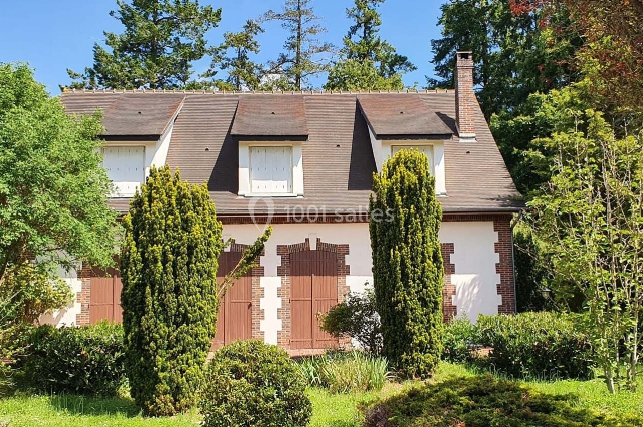 Maison traditionnelle avec toit en pente, volets marron et jardin arboré sous un ciel bleu.