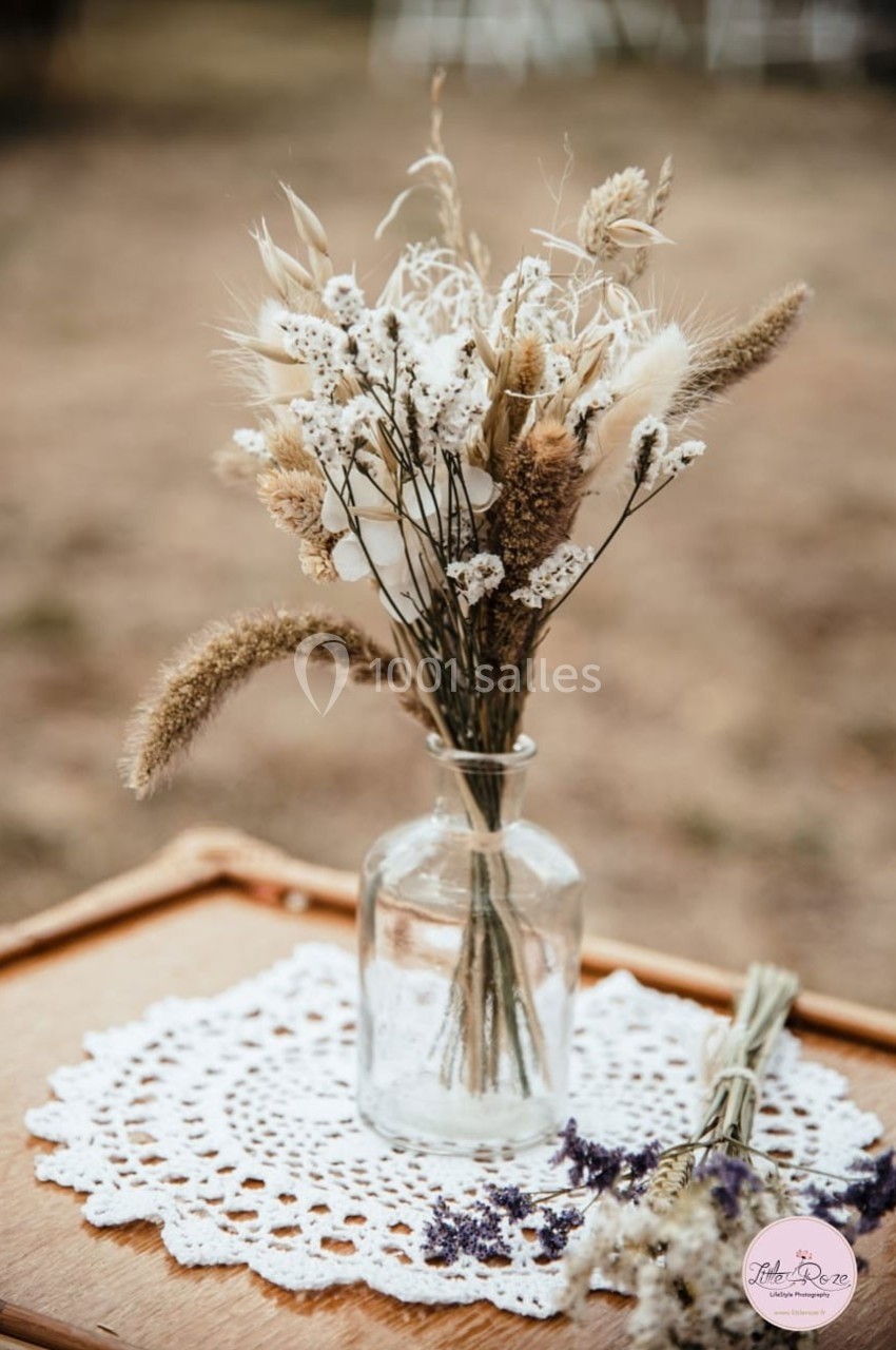 Bouquet de fleurs séchées dans un vase en verre, posé sur une nappe en dentelle sur une table en bois.