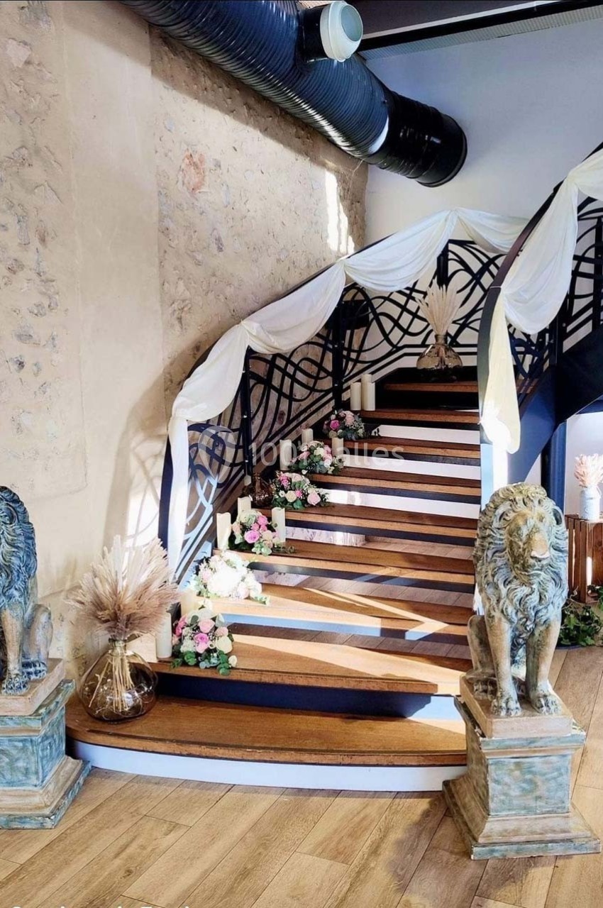 Escalier en bois décoré de fleurs et drapés blancs, encadré par deux statues de lions dans une salle au mur en pierre.