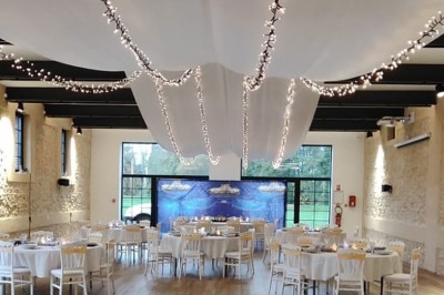 Salle de réception décorée avec des tables rondes, nappes blanches, chaises blanches et centres de table fleuris.