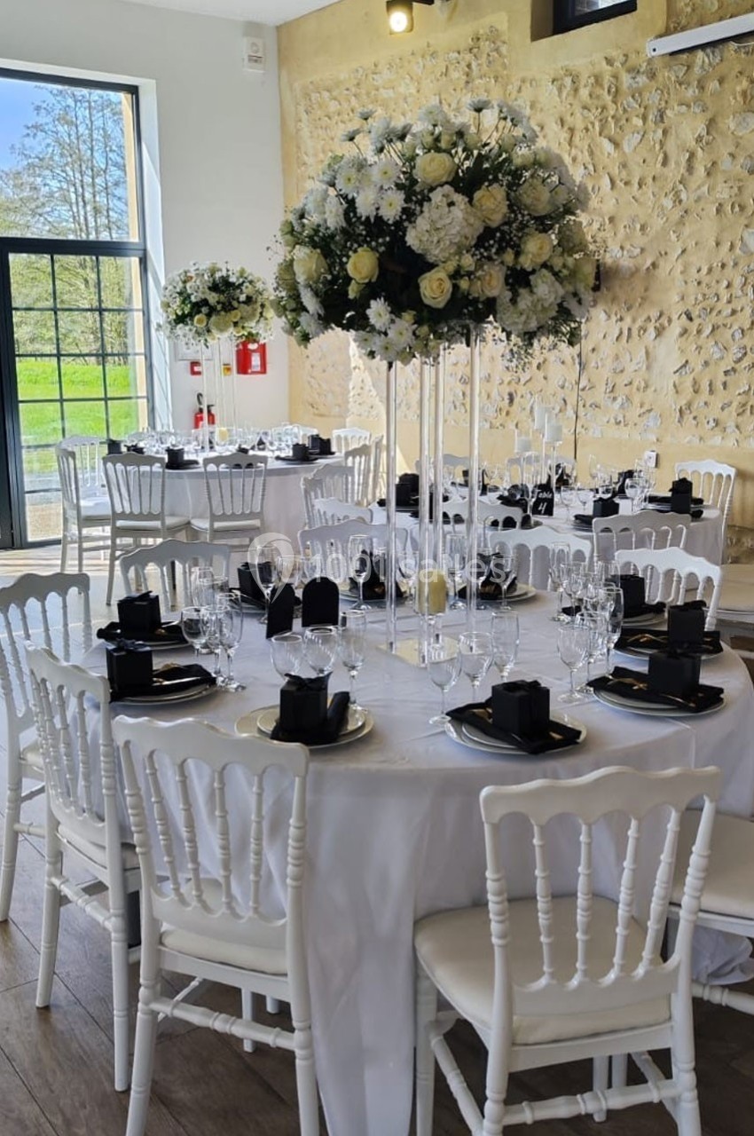 Salle de réception décorée avec des tables rondes, nappes blanches, chaises blanches et centres de table fleuris.