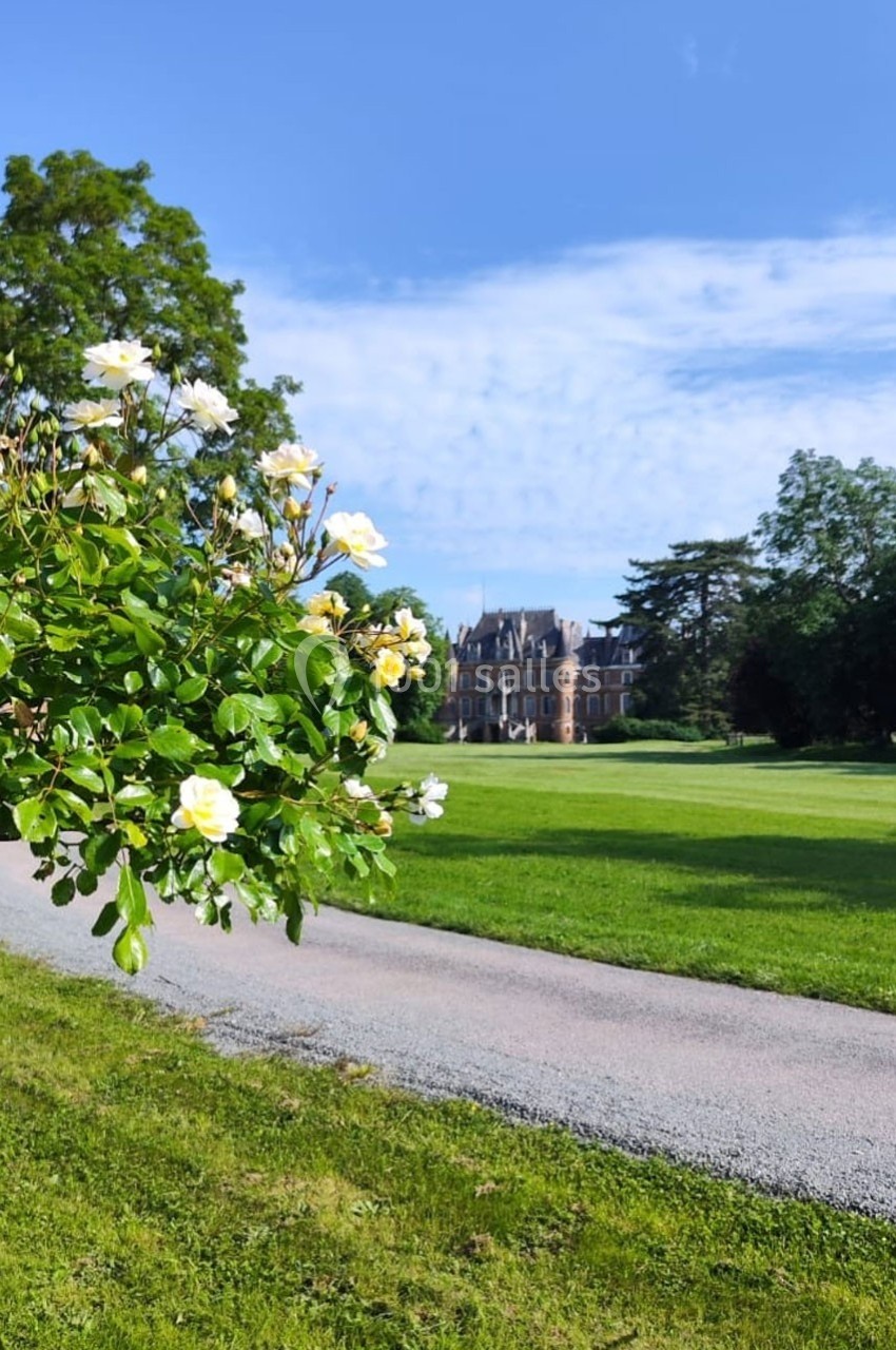 Allée bordée de pelouse avec des roses blanches au premier plan et un château entouré d'arbres en arrière-plan.