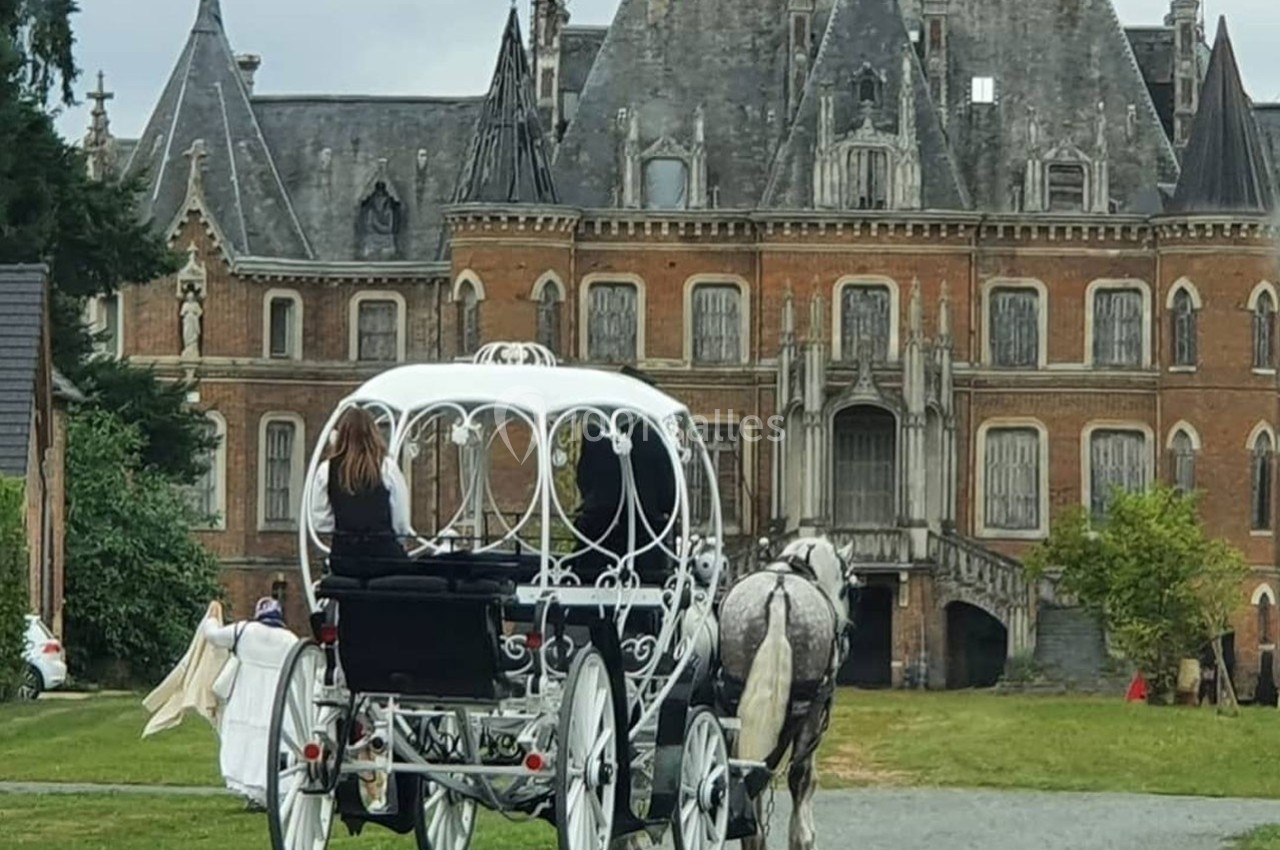 Une calèche blanche tirée par un cheval stationne devant un château en briques avec des toits en ardoise.