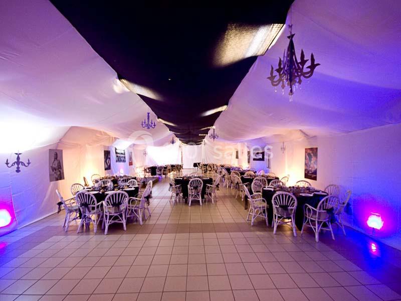Salle de réception décorée avec des tables rondes, chaises blanches et éclairage coloré dans une ambiance festive.